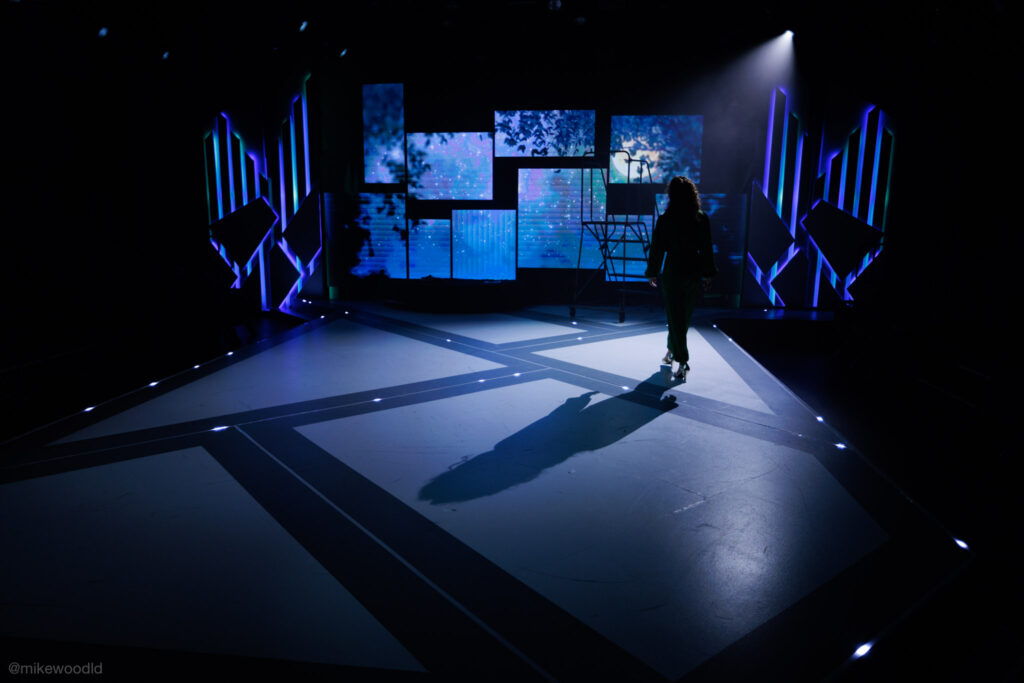 Tell Me On A Sunday — solo performer walking upstage, single wide beam casting a long shadow behind her
