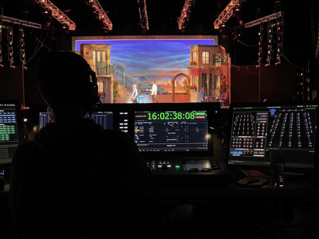An apprentice silhouetted at the tech table during a performance, consoles and timecode displays in the foreground, the lit set in the background.