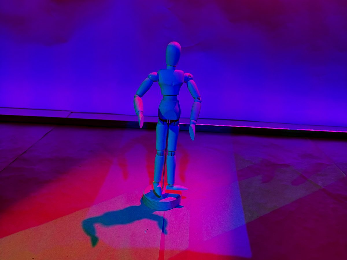 A wooden artist’s mannequin posed on a stage washed in saturated blue and magenta light, casting a long shadow.